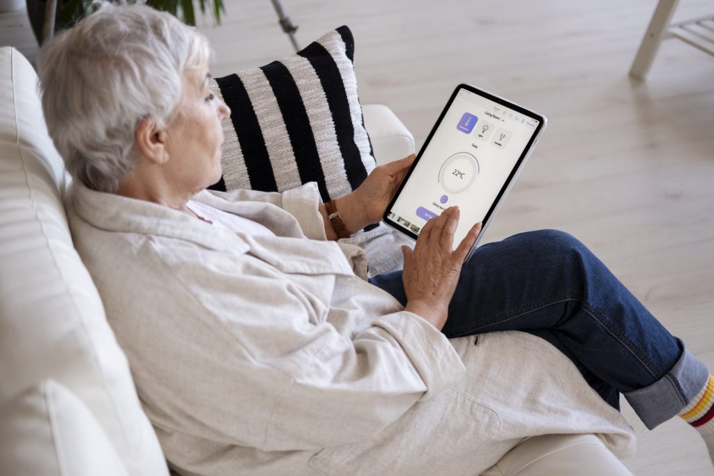 Elderly white woman with white short hair sitting in a white couch using a smart-home device to regulate the temperature. 