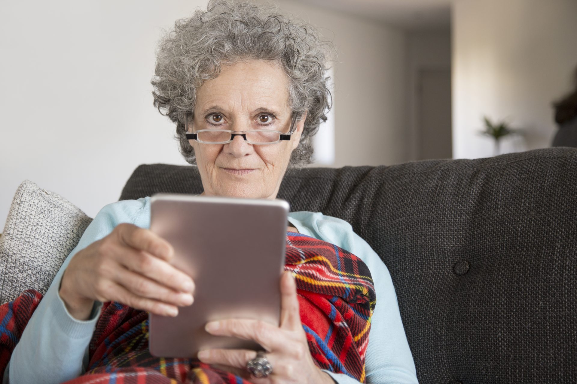 Older woman with curly hair and glasses seating in a dark grey couch using a modern device home.